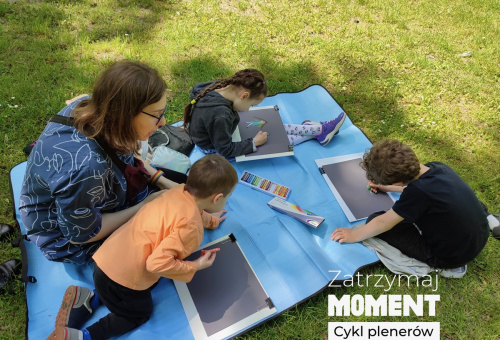 Organizatorka warsztatów siedzi z dziećmi na niebieskim kocu rozłożonym na jasnozielonym kobiercu kiełkującej wiosennej trawy. Dzieci rysują pastelami na szarych brystolach.