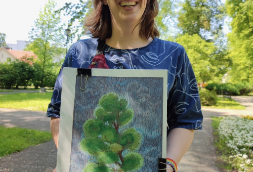 Młoda kobieta w okularach ubrana w granatową suknię trzyma pracę przedstawiającą drzewo. Jego ulistnione konary tworzą jajowato-podłużne kępy zieleni, a otaczają je falujące odcienie błękitnego nieba.