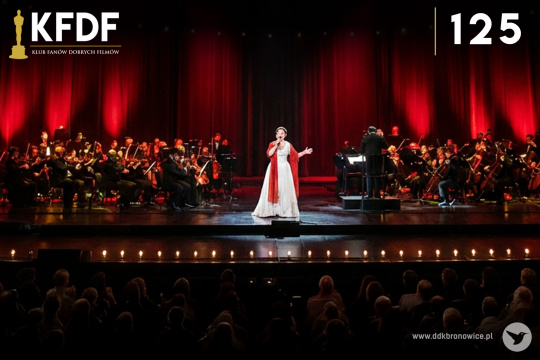 Grafika promująca wydarzenie cykliczne Klub Fanów Dobrych Filmów w Pracowniach Kultury Maki. Grafikę stanowi zdjęcie wielkiej sceny. W tle orkiestra i czerwone reflektory. Na środku sceny artystka w białej sukni. W lewym górnym rogu grafiki, mała grafika Oscara. Obok, po prawej stronie, biały napis: “KFDF”. Pod nim cienka żółta linia, a pod nią napis: “KLUB FANÓW DOBRYCH FILMÓW”. W prawym górnym rogu grafiki, cienka, żółta linia pionowa i obok biały napis “125”. W prawym dolnym rogu szary napis www.ddkbronowice.pl, obok napisu mały koliber na szarym tle.