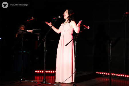 Kolorowe zdjęcie. Kobieta w sukni na scenie śpiewa do mikrofonu. Rozkłada swoje dłonie na boki.