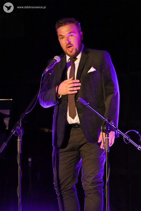 Kolorowe zdjęcie. Mężczyzna w garniturze i krawacie śpiewa do mikrofonu.