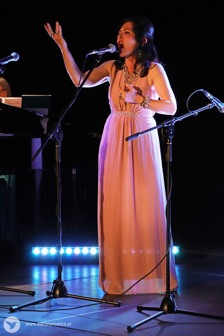 Kolorowe zdjęcie. Kobieta śpiewa na scenie z gestykulacją. Wyciąga prawą dłoń w górę.