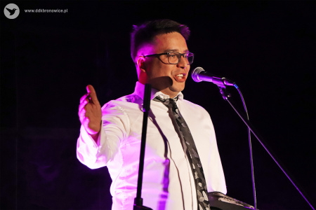 Kolorowe zdjęcie. Mężczyzna w białej koszuli i krawacie śpiewa do mikrofonu. Swoją prawą dłoń kieruje do prawego boku coś wskazując.