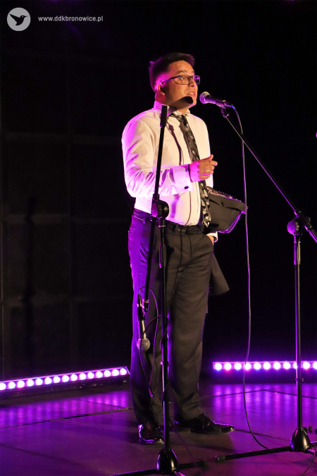 Kolorowe zdjęcie. Mężczyzna w białej koszuli i krawacie śpiewa do mikrofonu na statywie na scenie.