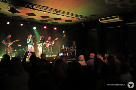 Kolorowe zdjęcie. Z daleka widać na scenie zespół muzyczny. Wokalistka śpiewa do mikrofonu, trzech gitarzystów gra na gitarach elektrycznych.