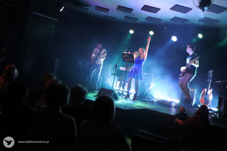 Kolorowe zdjęcie. Na scenie wokalistka śpiewa do mikrofonu i unosi swoją lewą rękę do góry. Obok niej dwóch gitarzystów gra na gitarach.