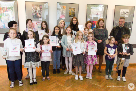 Kolorowe zdjęcie. Laureaci wystawy wraz z opiekunami i panią Dyrektor DDK Bronowice pozują do zdjęcia na tle wystawy. Dzieci trzymają w dłoniach dyplomy.