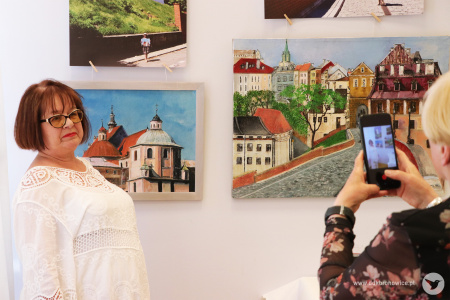 Kolorowe zdjęcie. Kobieta pozuje do zdjęcia przy obrazach z wystawy. Po prawej stronie widać telefon w dłoniach kobiety, która wykonuje zdjęcie.