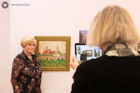 Kolorowe zdjęcie. Kobieta pozuje do zdjęcia przy obrazie przedstawiającym Lublin w złotej ramie. Po prawe stronie kobieta z tabletem robi jej zdjęcie.