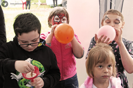 Kolorowe zdjęcie. Dzieci pod namiotem na dworze. Dwoje z tyłu z pomalowanymi twarzami dmucha balony. Z przodu chłopiec przesypuje przez lejek mąkę. Obok stoi dziewczynka.