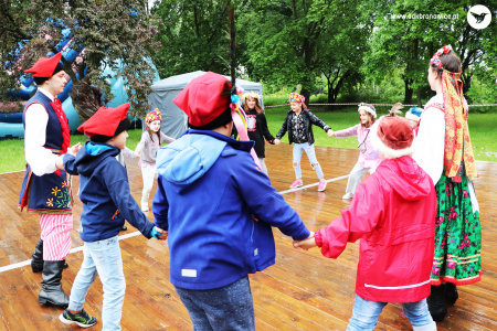 Kolorowe zdjęcie. Na dworze. Na drewnianym parkiecie dzieci w ludowych czapkach z parą w ludowych strojach tańczą w kole.