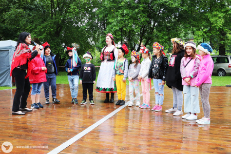 Kolorowe zdjęcie. Na dworze. Na drewnianym parkiecie. Grupa dzieci z kobietami w ludowych strojach śpiewają stojąc w półkolu.