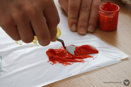 Kolorowe zdjęcie. Kobiece dłonie rozprowadzają szpachelką czerwoną farbę na kartce papieru w koszulce.