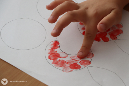 Kolorowe zdjęcie. Dziecięce palce malują czerwoną farbą naszkicowany ołówkiem półksiężyc na białej kartce papieru.