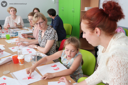 Kolorowe zdjęcie. Dziewczynka siedzi przy stole i moczy palec wskazujący w czerwonej farbie. Nad nią po prawej stronie stoi instruktorka. Po lewej stronie siedzą pozostałe uczestniczki warsztatów i tworzą swoje prace techniką petrykiwka.