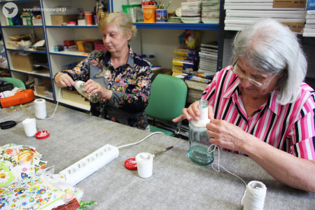 Kolorowe zdjęcie. Dwie kobiety siedzą przy stole i owijają sznurek wokół szklanych butelek.