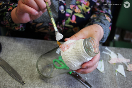 Kolorowe zdjęcie. Kobiece dłonie malują pędzelkiem po szklanej butelce z nawiniętym sznurkiem.