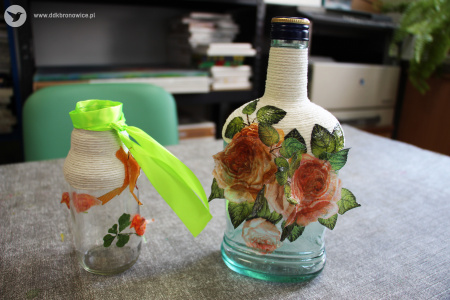 Kolorowe zdjęcie. Na stole stoją dwie szklane butelki. Butelka większa po prawej stronie jest przyozdobiona elementami róż oraz sznurkiem. Po lewej stoi mniejsza butelka owinięta zieloną wstążką, sznurkiem oraz ma naklejone elementy kwiatów.