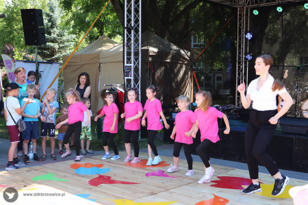 Kolorowe zdjęcie. Dziewczynki w różowych koszulkach i czarnych legginsach tańczą z instruktorką na podeście przed sceną.