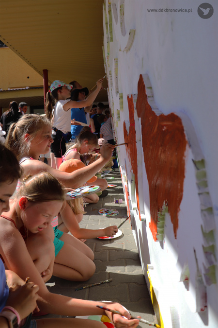 Kolorowe zdjęcie. Dzieci kucają przy chodniku i malują pędzlami wielkoformatową tablicę.