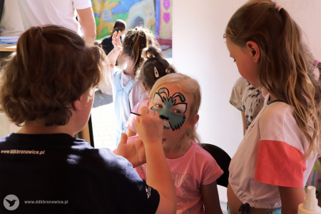 Kolorowe zdjęcie. Instruktorka DDK Bronowice maluje twarz dziewczynce. Obok stoją inne dziewczynki w kolejce.