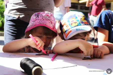 Kolorowe zdjęcie. Dziewczynka i chłopiec w czapkach malują flamastrami po drewnianych zawieszkach przy stoliku.