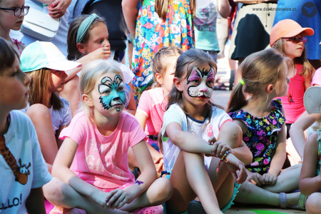 Kolorowe zdjęcie. Dziewczynki z pomalowanymi twarzami siedzą na chodniku. Wokół nich siedzą w tle inne dzieci.