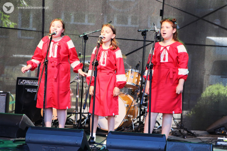 Kolorowe zdjęcie. Trzy dziewczynki w czerwonych sukienkach śpiewają do mikrofonów na scenie.