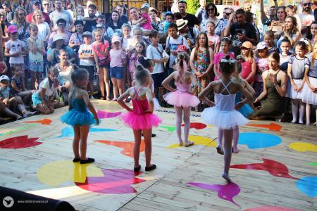 Kolorowe zdjęcie. Grupa dziewczynek w strojach baletnic i tiulowych spódniczkach tańczy na podeście tyłem do fotografa. Wokół podestu stoi licznie zgromadzona publiczność.