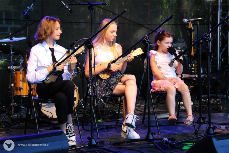 Kolorowe zdjęcie. Na scenie dwójka młodych ludzi  siedzi na krzesłach i gra na ukulele. Obok nich na krześle siedzi dziewczynka i śpiewa do mikrofonu.