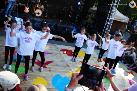 Kolorowe zdjęcie. Grupa dziewczynek w koszulkach z napisem Groovy Kids i czarnych legginsach tańczy na podeście przed sceną.
