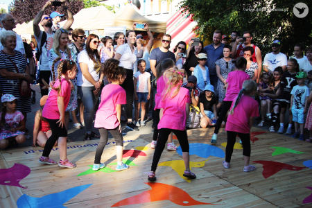 Kolorowe zdjęcie. Grupa dziewczynek w różowych koszulkach i czarnych legginsach tańczy na podeście tyłem do fotografa. Wokół zgromadzona jest licznie publiczność.