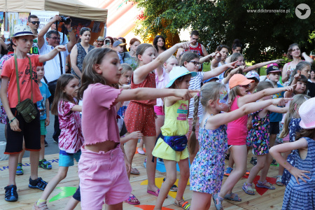 Kolorowe zdjęcie. Na dworze. Grupa dzieci tańczy na podeście. Prawą dłoń prostują przed siebie i wskazują na coś palcem.