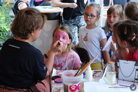 Kolorowe zdjęcie. Instruktorka DDK Bronowice maluje twarz dziewczynce. Za dziewczynką stoi kolejka dzieci.