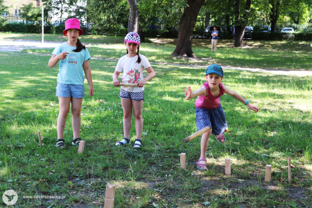 Kolorowe zdjęcie. Trzy dziewczynki grają na trawie w grę króla z użyciem drewnianych bloczków.