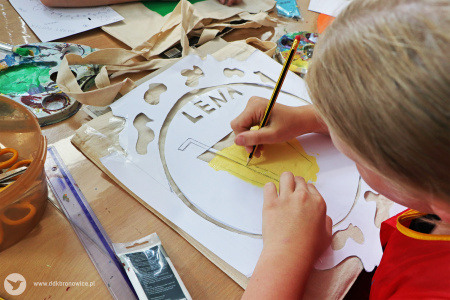 Kolorowe zdjęcie. Dziecięca dłoń odrysowuje ołówkiem z szablonu szkic szklanki ze słomką i napisem LENA na bawełnianej torbie.