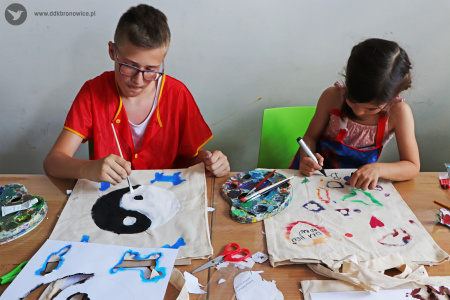 Kolorowe zdjęcie. Chłopiec i dziewczynka siedzą przy stoliku i malują farbami na bawełnianych torbach.