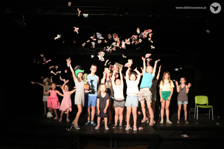 Kolorowe zdjęcie. Grupa dzieci pozuje do zdjęcia na scenie. Podrzucają do góry kawałki kolorowej gazety. Cieszą się.