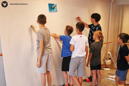 Kolorowe zdjęcie. Chłopcy stoją przy wielkoformatowej tablicy i szkicują na niej rekina.