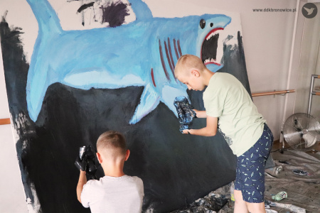 Kolorowe zdjęcie. Dwóch chłopców maluje farbami rekina na wielkoformatowej tablicy.