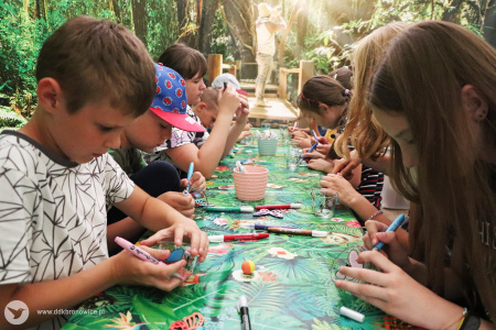 Kolorowe zdjęcie. Dzieci siedzą przy stole. Malują po szklanych słoiczkach flamastrami.