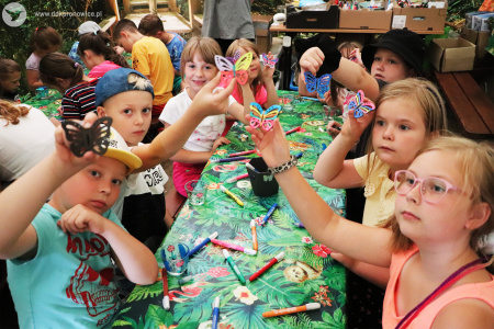 Kolorowe zdjęcie. Dzieci siedzą przy stoliku. Unoszą do góry ręce, w których trzymają pomalowane flamastrami motyle.