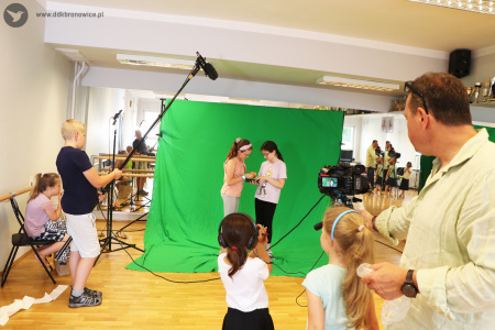 Kolorowe zdjęcie. Grupa dzieci uczestniczy w warsztatach filmowych. Na środku stoi zielona ścianka. Na ściance stoją dwie dziewczynki bokiem. Po lewej stronie stoi chłopiec i trzyma na statywie mikrofon. Obok na krześle siedzi dziewczynka. Po prawej stronie stoi instruktor z dwiema dziewczynkami za kamerą.