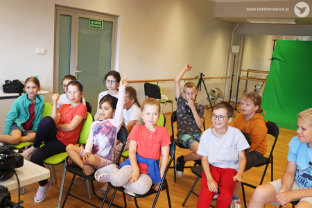 Kolorowe zdjęcie. Dzieci siedzą na krzesłach przodem do fotografa. Dwoje z nich unosi ręce do góry.