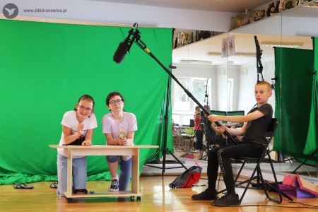 Kolorowe zdjęcie. Dwie dziewczynki klęczą za stolikiem na tle zielonej ścianki. Po prawej stronie na krześle siedzi chłopiec i trzyma nad dziewczynkami mikrofon na statywie.