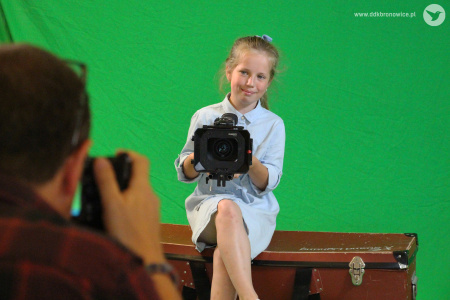 Kolorowe zdjęcie. Dziewczynka siedzi na skrzyni z kamerą w dłoniach i uśmiecha się. Za nią widać zieloną ścianę. Na pierwszym planie po lewej stronie widać rozmazaną sylwetkę instruktora, który nagrywa dziewczynkę.