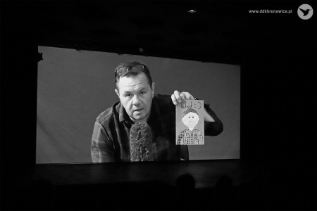 Czarno-białe zdjęcie. Ekran kinowy. Na ekranie postać instruktora mówi do mikrofonu i trzyma w lewej dłoni rysunek z chłopcem i napisem Michał.