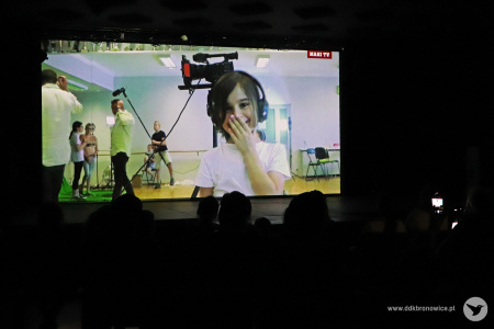 Kolorowe zdjęcie. Ekran w sali widowiskowej. Na ekranie prezentowany jest film z warsztatów filmowych.