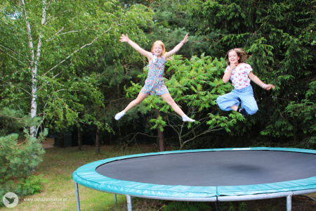 Kolorowe zdjęcie. Na dworze. Wielka trampolina. Dwie dziewczynki skaczą na trampolinie.