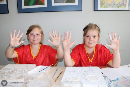 Kolorowe zdjęcie. Dwie dziewczynki siedzą przy stoliku. Unoszą wybrudzone w gipsie ręce w kierunku fotografa. Uśmiechają się.